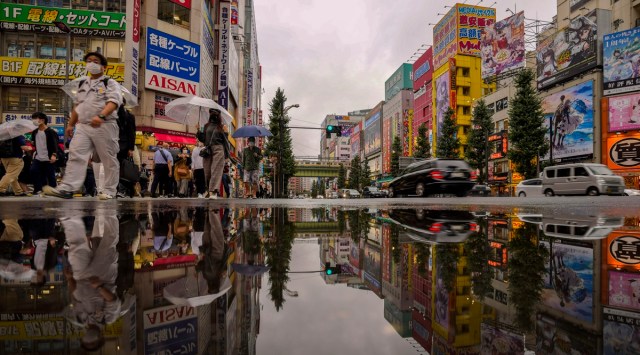 People wearing protective masks are reflected in a puddle as they walk across a street in a shopping district on Oct. 12, 2021, in Tokyo. (AP)