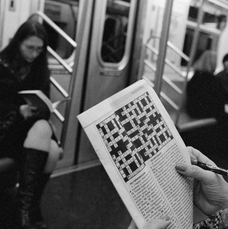 In a metro train coach, a woman holds a newspaper showing a crossword she is solving.  The photo shows her hand and the newspaper in focus, with other passengers in the blurry background.