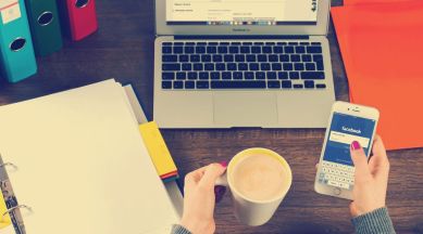 A person's hands are visilbe. One holds coffee while the other holds a phone with Facebook on the screen. There is a desk in the background with a laptop on it.