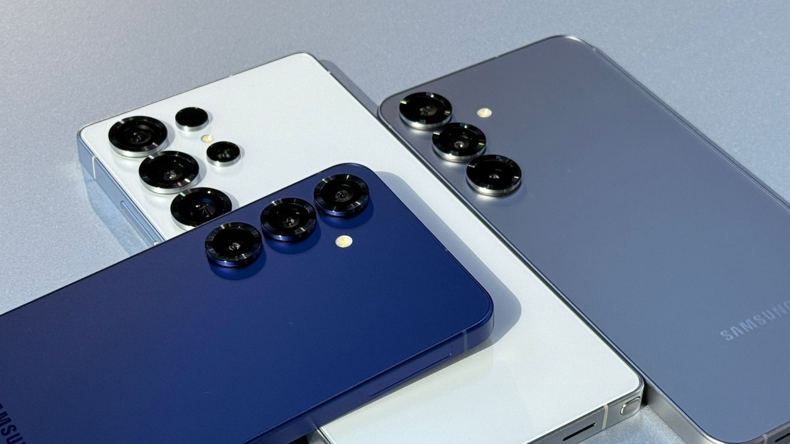 The image shows a close-up view of the backs of three modern smartphones arranged overlapping each other on a flat surface. One phone is white and features four camera lenses grouped together along with a flash, while the other two phones, one in a deep navy blue and the other in gray, each have three camera lenses and a flash in a similar configuration. The phones have a sleek, rectangular design with slightly rounded corners, and the Samsung logo is faintly visible on the gray phone. The lighting highlights the smooth finish and raised camera modules on each device.