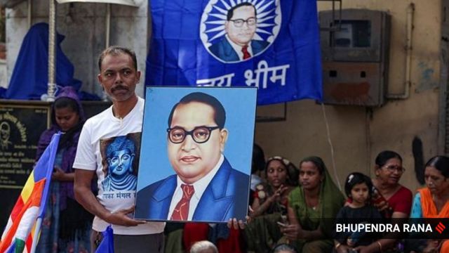 man holding poster of BR Ambedkar stands with a blue Ambedkar poster and other people sitting in the background