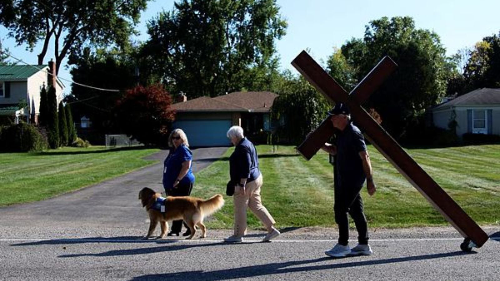 Dan Beazley, of Northville, carries a homemade cross past a therapy dog to The Church of Jesus Christ of Latter-day Saints in Grand Blanc Township, Mich., Tuesday, Sept. 30, 2025, after a man rammed his vehicle into the building before opening fire and setting the building ablaze Sunday morning. (AP Photo/Ryan Sun)