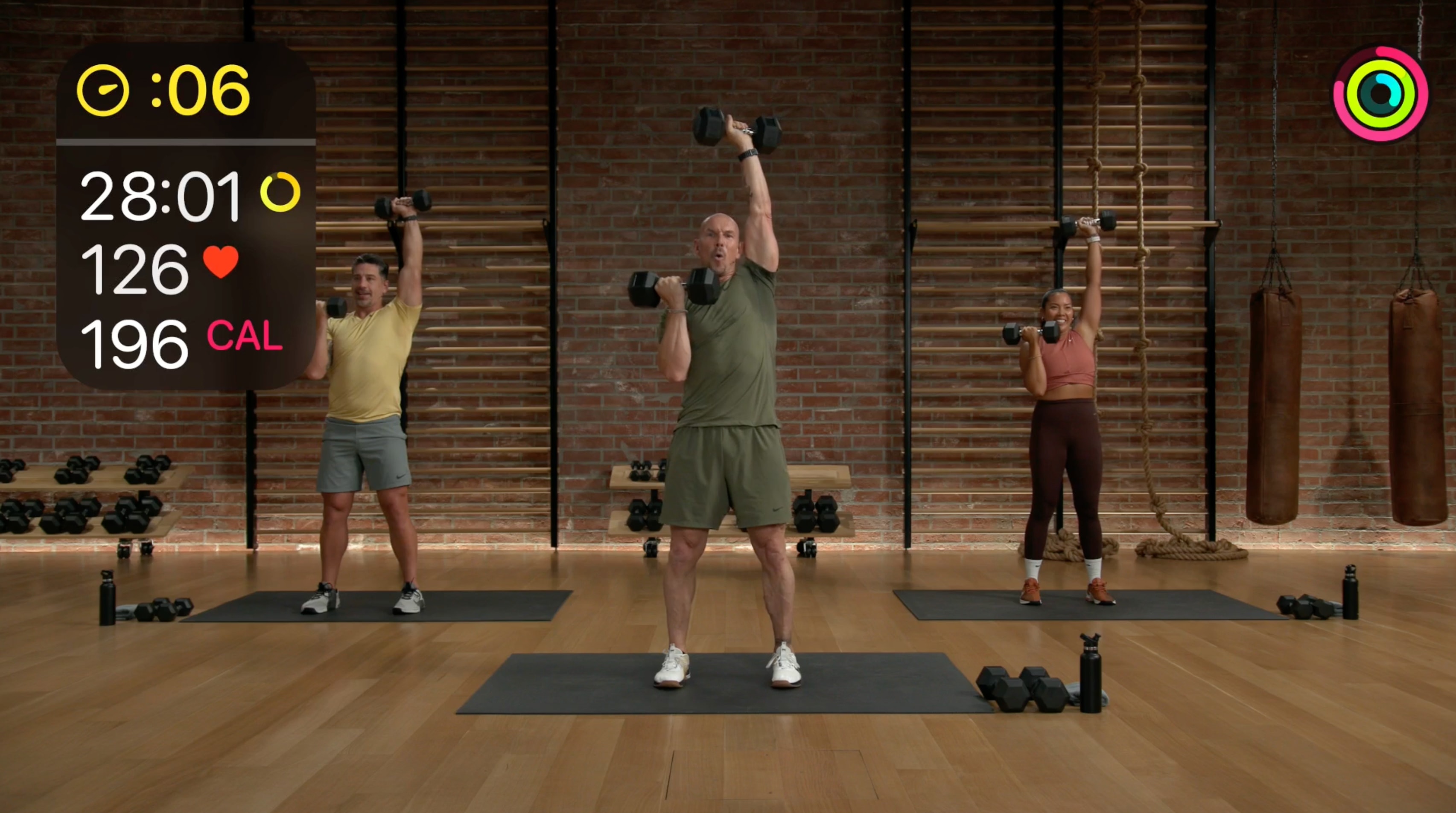 A strength training session being filmed in the Apple Fitness+ studio with real-time Apple Watch metrics on screen.
