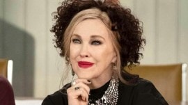 Catherine O'Hara in the costumery of Schitt's Creek's lead character Moira Rose. She is wearing a black wig on top of her blond hair, silver necklaces and has her hand under her chin.