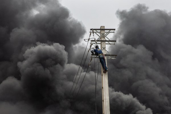 A worker stands on an electrical pole as plumes of smoke rise from an oil storage facility following overnight attacks by U.S. and Israeli forces in Tehran, Iran, Sunday, March 8, 2026. Most ordinary Iranians are cut off from the Internet, and for some it has been infuriating to have so little information about what is happening. (Credits: Arash Khamooshi)