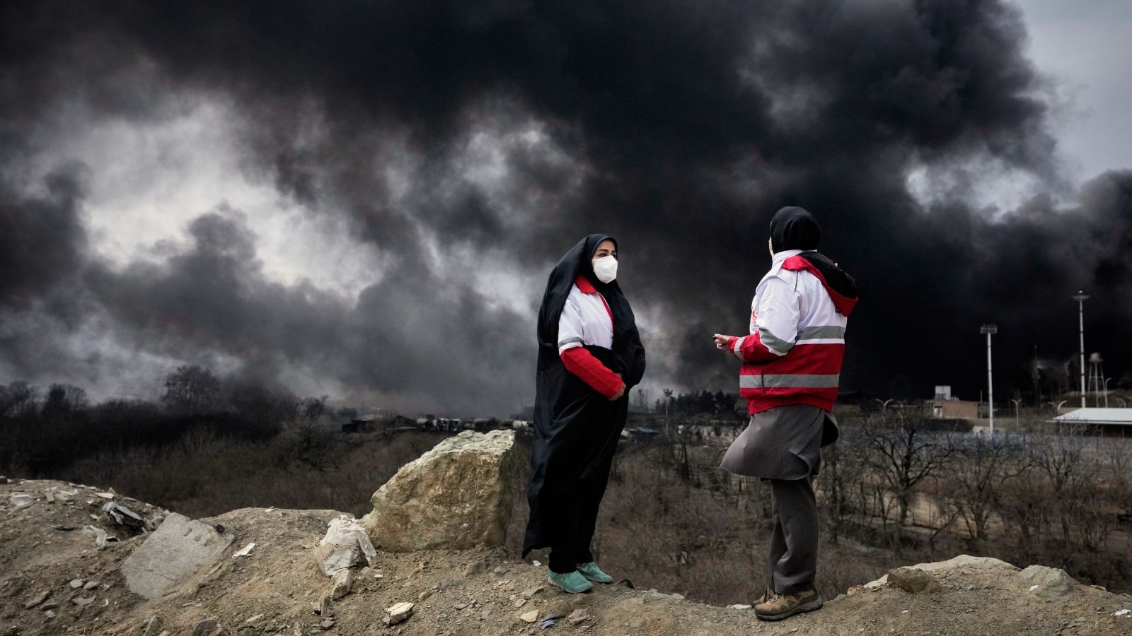 Two women from the Iranian Red Crescent Society stand as a thick plume of smoke from a U.S.-Israeli strike on an oil storage facility late Saturday rises into the sky in Tehran