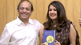 A photo of author Vikram Seth in a white shirt, with translator Mohini Gupta in a saree standing together at a book launch. They are holding copies of the new bilingual book, ‘Idhar Udhar ke Jangli Kisse,’ and smiling warmly. The background is a stage with event signage.