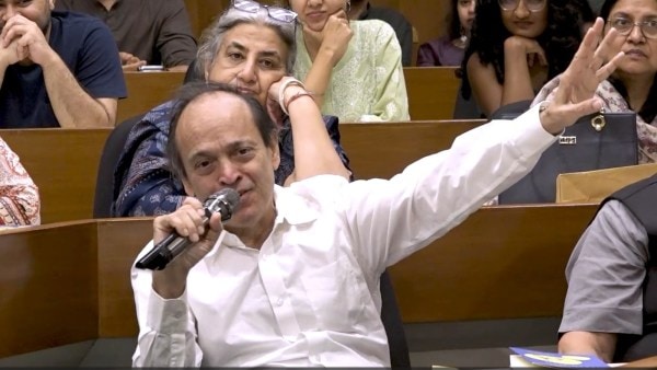 A candid photograph of author Vikram Seth speaking into a microphone during a literary event. He is wearing a white button-down shirt and gesturing with his left arm raised as he addresses an audience seated in a tiered lecture hall. Behind him, several attendees are seen listening intently. The lighting is warm and natural, capturing a moment of active intellectual exchange.
