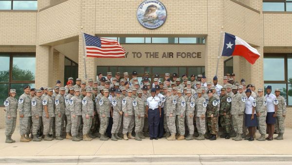 A large group of uniformed U.S. Air Force personnel stand outside a training wing building at Joint Base San Antonio-Lackland in Texas, with American and Texas state flags displayed overhead.