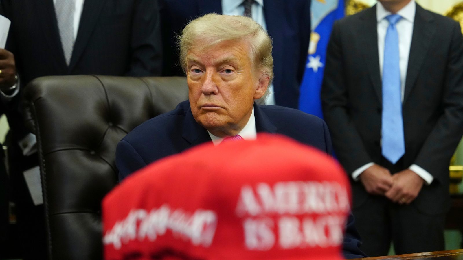 President Donald Trump sits in the Oval Office of the White House alongside a stack of red caps stating "America is Back" (AP Photo)