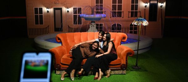 Visitors pose on the iconic orange couch at the FRIENDS exhibition in Mumbai, with a fountain-inspired backdrop recreating the show's opening scene. (Express Photo by Sankhadeep Banerjee)