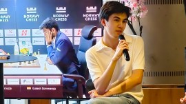 World championship contender Javokhir Sindarov speaks at an event while a TV behind shows a live game between world champion Gukesh and Alireza Firouzja at Norway Chess 2026. (Express Photo by Amit Kamath)