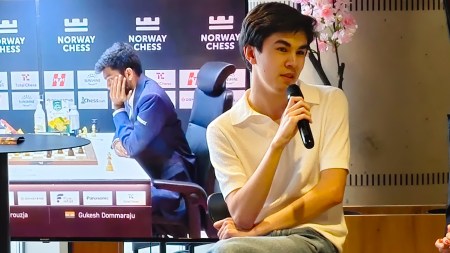 World championship contender Javokhir Sindarov speaks at an event while a TV behind shows a live game between world champion Gukesh and Alireza Firouzja at Norway Chess 2026. (Express Photo by Amit Kamath)