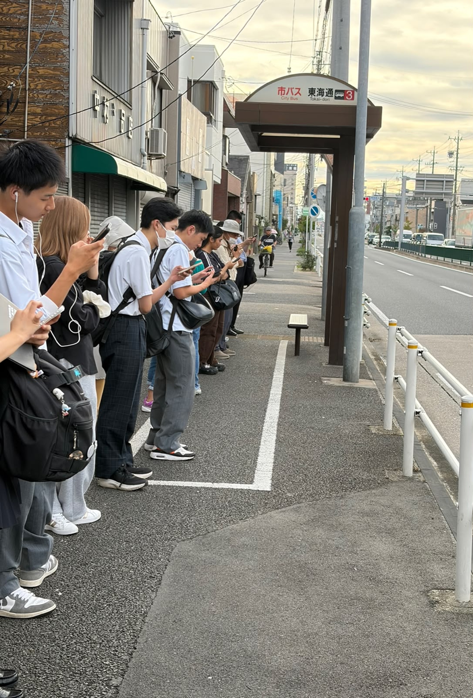 Heads are bowed, thumbs flicking, faces lit by the glow of phone screens. A few feet away, people are waiting together, yet each seems to be somewhere else.